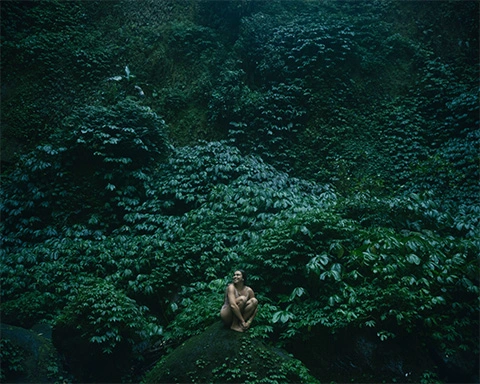 Girl in a swim suit sitting on a rock with a cliff of dark green leaves behind her
