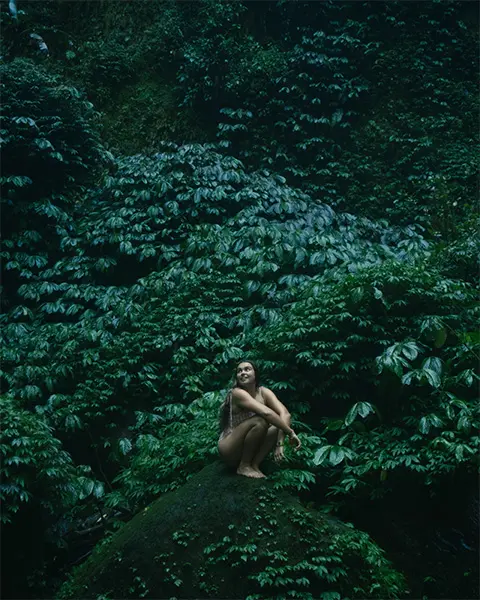 Girl in a swim suit sitting on a rock with a cliff of dark green leaves behind her