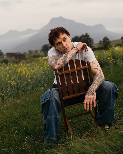 Man sitting on a wooden chair in tall grass with canola and mountains in the background