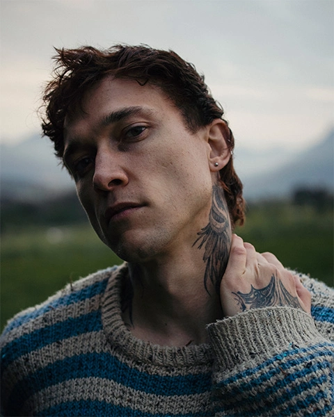 Portrait of a man with tattoos in a field holding his neck while wearing a wool sweater
