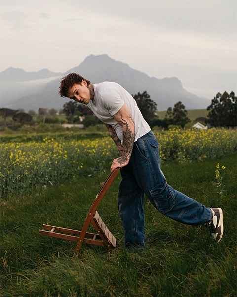 Man leaning on a wooden chair in tall grass with canola and mountains in the background