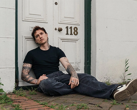 Man sitting against a white door with number 118 in the street wearing a dark jean and black shirt