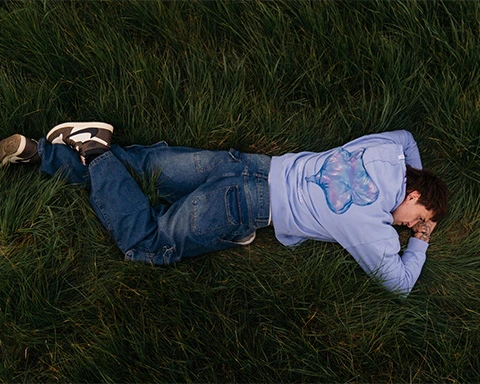 Man lying in tall green grass on his stomach wearing a blue jean and purple hoodie