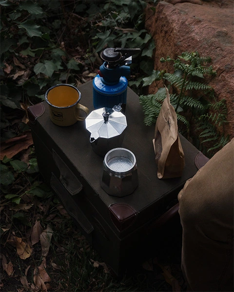 Gas stove, bialette coffee maker, a tin cup and Mannabrew packet on a crate in the woods