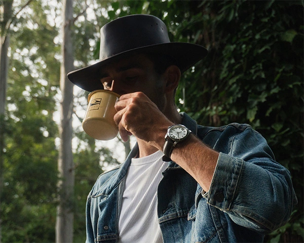 Man wearing a denim jacket and leather hat drinking Mannabrew in the woods
